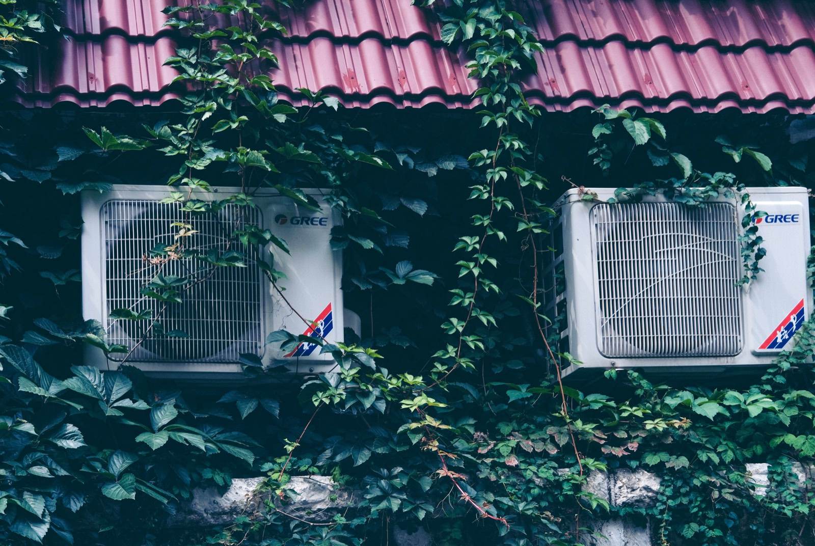 Two Gree outdoor condenser units mounted on an ivy-covered wall beneath a tiled roof